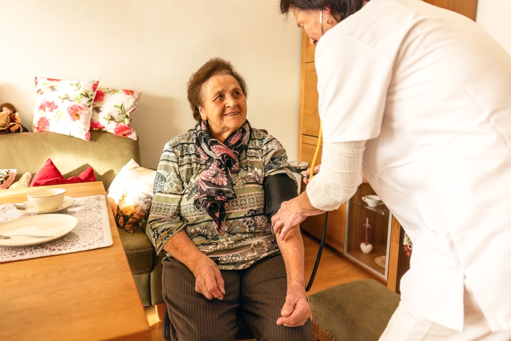 Licensed nurse providing skilled nursing care to an elderly patient at home, checking vital signs and reviewing medical treatment plan.